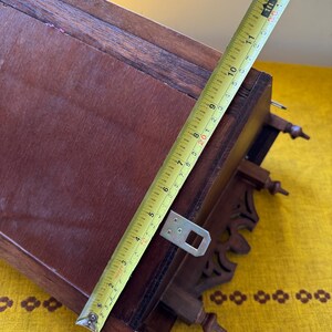 May include: A dark brown wooden wall-mounted shelf featuring decorative cut-out details and three small finials at the base. A yellow measuring tape is placed diagonally across the shelf, showing a length of approximately 13 inches.