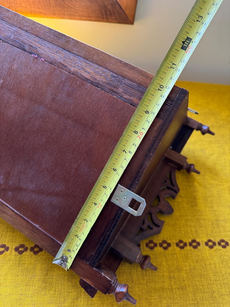 May include: A dark brown wooden wall-mounted shelf featuring decorative cut-out details and three small finials at the base. A yellow measuring tape is placed diagonally across the shelf, showing a length of approximately 13 inches.