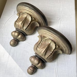 May include: Two decorative wall sconces with a gold and bronze finish. Each sconce features a stylized leaf design and a rounded shelf. The sconces are made of a sturdy material and are designed to hold small items.
