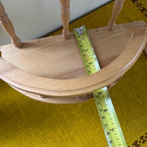 May include: A light-colored wooden shelf with a curved front and spindle legs. A yellow measuring tape is placed across the shelf, showing measurements in inches. The shelf appears to be unfinished.