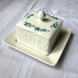 May include: A square, off-white ceramic butter dish with a lid, set on a matching rectangular base. The lid features a floral design in teal and a small, rounded handle. The dish has a textured basketweave pattern.