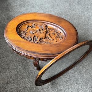 May include: Oval wooden side table with a detailed carved relief of cherubs and floral elements. The table has a dark brown finish and ornate legs. A detached oval frame rests beside the table.