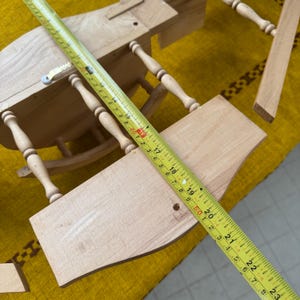 May include: A partially assembled wooden dollhouse with a yellow measuring tape laid across the pieces. The tape shows measurements in inches. The dollhouse has a table and chair design, with turned legs and a curved seat.