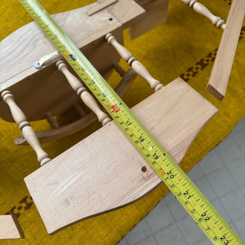 May include: A partially assembled wooden dollhouse with a yellow measuring tape laid across the pieces. The tape shows measurements in inches. The dollhouse has a table and chair design, with turned legs and a curved seat.