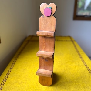 May include: A wooden shelf with three tiers and a heart-shaped top. A pink apple with a green leaf is attached to the heart. The shelf is made of light brown wood and is designed to hold small items.