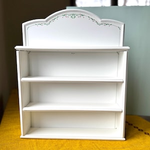 May include: A white, three-tiered wall shelf with a decorative top. The top features a curved design with a green and red floral pattern. The shelves are open and ready for display. The shelf is made of wood.