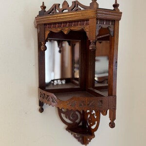 May include: A dark brown wooden corner wall shelf featuring two tiers, mirrored back panels, and intricate carved detailing along the top and bottom edges. The structure includes decorative finials and a cutout pattern on the lower shelf apron.