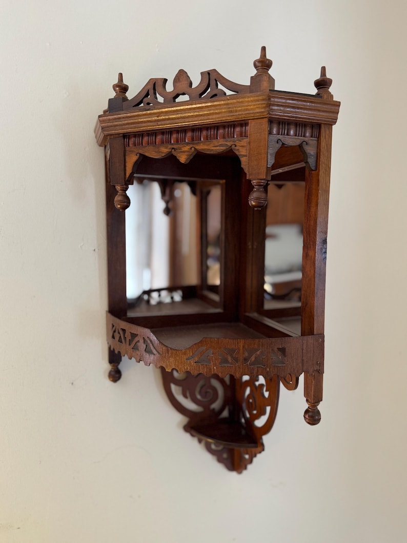 May include: A dark brown wooden corner wall shelf featuring two tiers, mirrored back panels, and intricate carved detailing along the top and bottom edges. The structure includes decorative finials and a cutout pattern on the lower shelf apron.