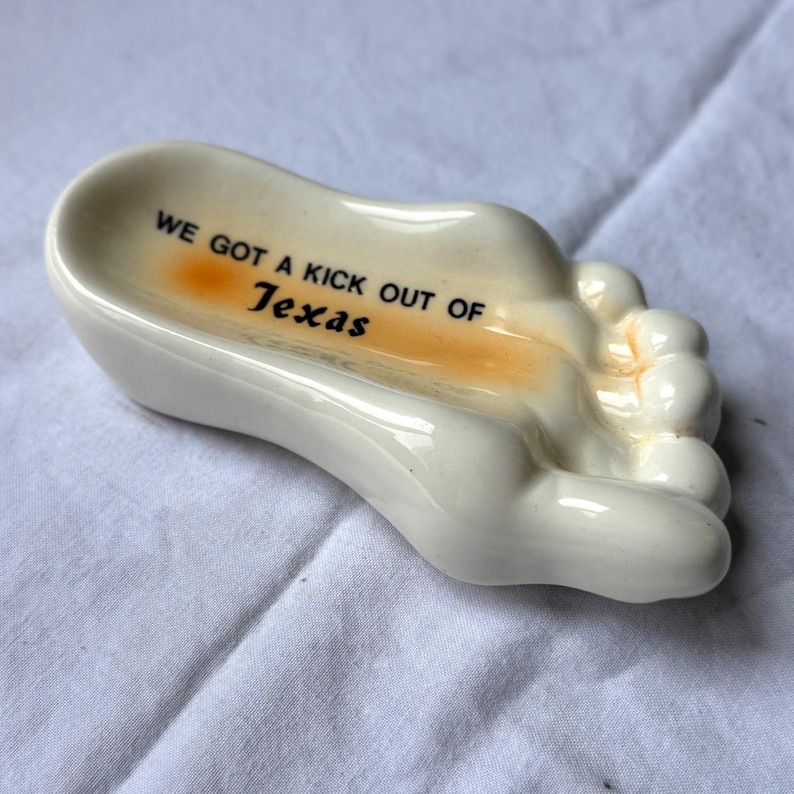 May include: A ceramic foot-shaped ashtray in off-white with the text "WE GOT A KICK OUT OF Texas" in black. The inside of the ashtray has an orange-yellow hue. The ashtray is on a white surface.