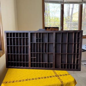 May include: A dark brown wooden antique printer's tray with numerous compartments of different sizes. The rectangular tray rests on a yellow tablecloth. The word "Antiques" is visible in the background.