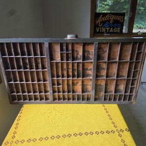 May include: A rectangular wooden printer's type drawer featuring numerous small, varied compartments with a dark exterior frame and worn, light brown interior wood surfaces. A single metal handle is centered on the top edge of the frame.