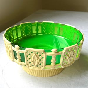 May include: A decorative bowl with a vibrant green interior and a cream-colored, lattice-style exterior. The bowl sits on a cream-colored pedestal base, creating a layered effect. The design features a repeating pattern around the rim.