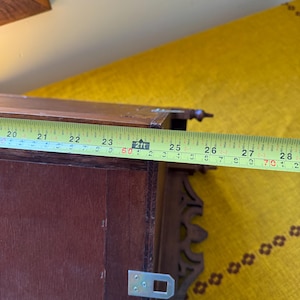 May include: A dark brown wooden wall shelf featuring decorative carved side brackets and metal mounting hardware. A yellow measuring tape is positioned across the top, indicating a width of approximately 8.5 inches for this specific section.