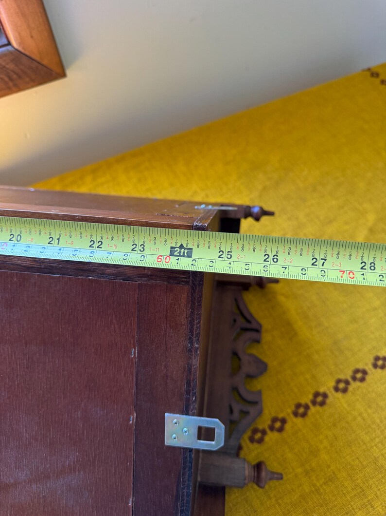 May include: A dark brown wooden wall shelf featuring decorative carved side brackets and metal mounting hardware. A yellow measuring tape is positioned across the top, indicating a width of approximately 8.5 inches for this specific section.