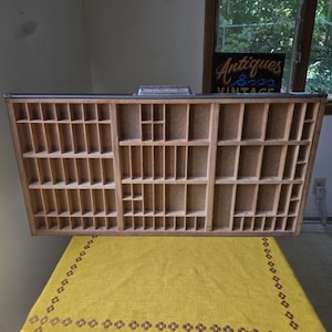 May include: A rectangular wooden printer's drawer featuring numerous small, varied compartments arranged in a grid pattern. The light-coloured wood has a natural finish, and a metal handle is attached to the top edge of the frame.