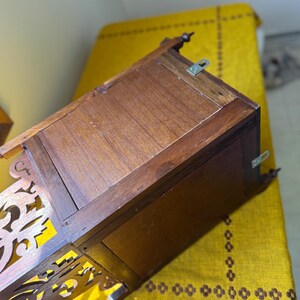 May include: A brown wooden wall-mounted box with a rectangular frame and a decorative cutout panel on the side. The back panel features two metal mounting brackets attached to the top and side edges, showing a natural wood grain finish throughout.