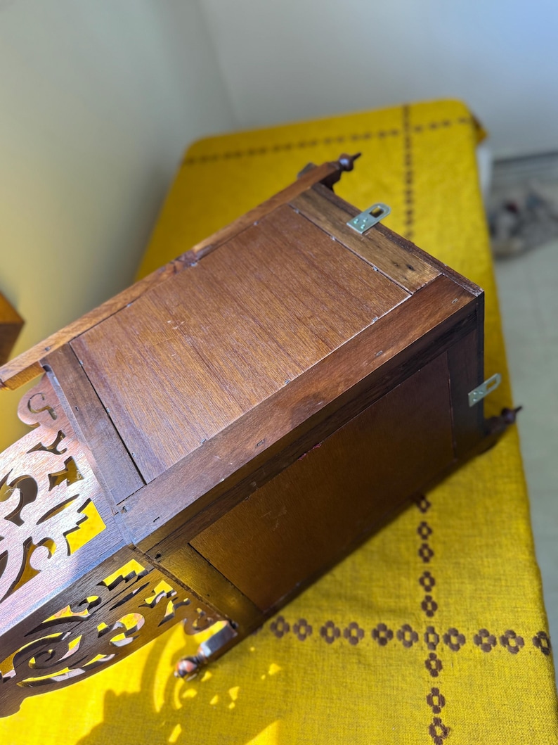 May include: A brown wooden wall-mounted box with a rectangular frame and a decorative cutout panel on the side. The back panel features two metal mounting brackets attached to the top and side edges, showing a natural wood grain finish throughout.