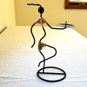 May include: Black metal candle holder in the shape of a stylized figure. The figure has a round head, woven tan accents, and holds a small, round candle plate. The base is a circular design.