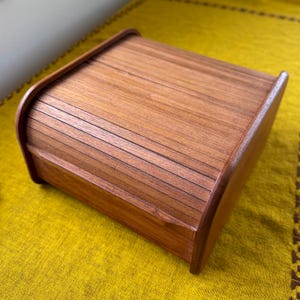 May include: A rectangular wooden bread box with a roll-top lid. The box is made of light brown wood with a visible grain. The lid curves smoothly over the top, and the box sits on a yellow surface.