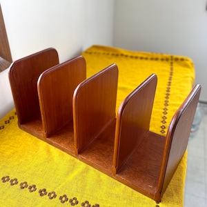 May include: A vintage wooden desk organizer with four vertical dividers. The organizer is made of a warm-toned wood and sits on a yellow patterned surface. The dividers have rounded tops and are evenly spaced.