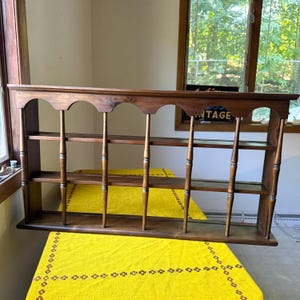 May include: A brown wooden wall shelf featuring three horizontal tiers and a decorative scalloped top edge. The structure is supported by six vertical turned spindles that connect the shelves, creating an open display unit with a warm wood finish.