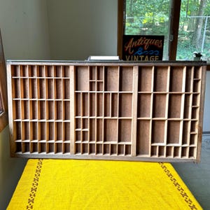 May include: A rectangular wooden printer's drawer featuring a grid of numerous small, open compartments of varying sizes. The frame is dark brown, and the interior dividers are a lighter wood tone. A metal handle is attached to the top center edge.