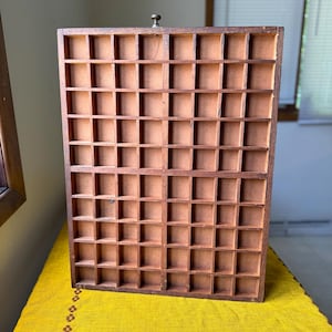 May include: A rectangular wooden display case featuring a grid of numerous small, square compartments. The frame and dividers are constructed from medium-toned brown wood, and a single small metal knob is centered at the top edge of the frame.