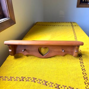 May include: A light brown wooden wall shelf featuring a flat top surface and a decorative front panel with a central heart-shaped cutout. The shelf includes two small wooden pegs extending from the front panel for hanging items.