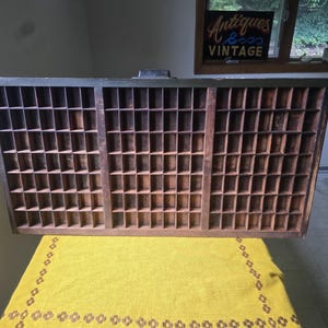 May include: A rectangular wooden printer's tray featuring a dark brown finish and numerous small, square compartments arranged in a grid pattern. The tray has a black metal handle centered on the top edge and a weathered, aged appearance.