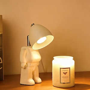 May include: A white, figure-shaped desk lamp with a pivoting head, casting a warm light. Next to it, a lit candle in a glass jar with a label. The lamp and candle are on a wooden surface, creating a cosy atmosphere.