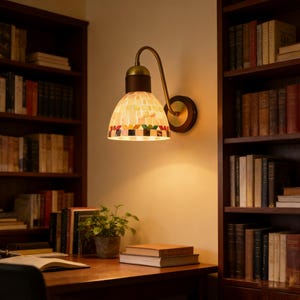 Puede incluir: Una lámpara de pared con una pantalla de mosaico de vidrio en un entorno de biblioteca. La lámpara tiene un brazo de latón y una base de madera. La pantalla está compuesta por pequeñas baldosas de colores. El fondo incluye estanterías llenas de libros y un escritorio de madera.