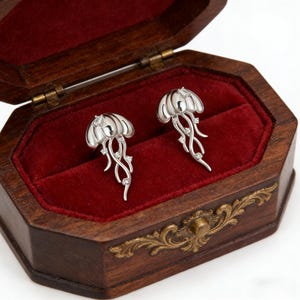 Op de afbeelding: Zilverkleurige oorringen in de vorm van kwallen met heldere edelsteenaccenten, gepresenteerd in een open houten sieradendoos met een rode fluwelen binnenkant. De doos heeft een decoratieve messing sluiting.