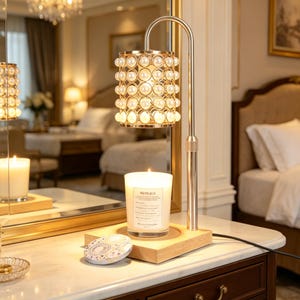 Puede incluir: Una lámpara decorativa dorada con una pantalla cilíndrica adornada con perlas falsas. La lámpara está sobre una base de madera, junto a una vela encendida en un tarro de cristal. También hay un pequeño recipiente redondo.