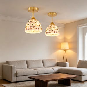 Könnte beinhalten: Zwei Pendelleuchten mit goldfarbenen Halterungen und weißen Schirmen. Die Schirme sind mit bunten, mosaikartigen Mustern verziert. Die Leuchten hängen von der Decke und beleuchten ein Wohnzimmer mit Sofa und Couchtisch.