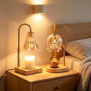 May include: Two decorative lamps with amber glass shades and gold-colored metal frames sit on a wooden nightstand. One lamp has a pleated shade, and the other has a flower-shaped shade. A lit candle and a jar candle are beneath the lamps.
