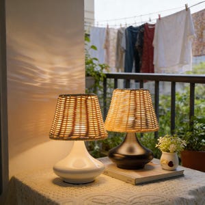 May include: Two decorative table lamps with woven rattan shades and curved bases. One lamp is white, the other is dark gray. The lamps sit on a table with a white lace tablecloth. A book and small vase are also on the table.