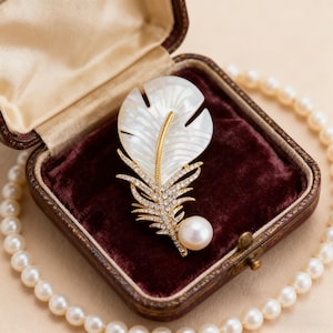 May include: A feather-shaped brooch with a pearl accent. The brooch features a white, iridescent feather design with a gold-colored stem and is encrusted with small, clear crystals. It is presented in a vintage-style jewelry box with a maroon velvet interior.