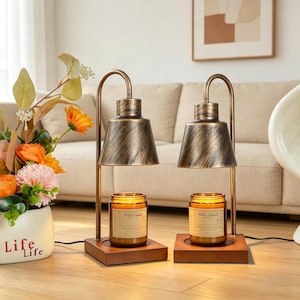 Puede incluir: Dos calentadores de velas con pantallas de color bronce y bases de madera. Cada calentador contiene una vela en tarro con una etiqueta. Los calentadores están sobre un suelo de madera, con un jarrón de flores y un sofá al fondo.