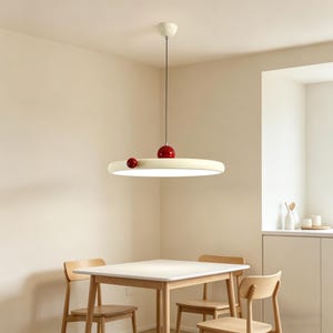 May include: A contemporary pendant light with a circular, cream-coloured shade and integrated LED lighting. Two red spheres are attached to the shade. The light hangs from a black cord and is fixed to the ceiling with a white canopy. Suitable for a dining area.