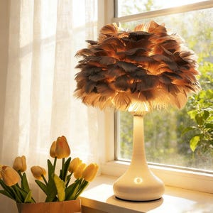 May include: A decorative table lamp with a white base and a shade made of brown and tan feathers. The lamp is turned on, emitting a warm glow. Yellow tulips in a brown paper vase sit on the windowsill.