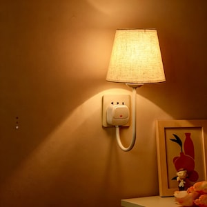 Może przedstawiać: Lampa ścienna z beżowym abażurem i białym, zakrzywionym ramieniem. Lampa jest podłączona do gniazdka ściennego. Ściana jest pomalowana na ciepły, neutralny kolor i widoczny jest obraz w ramce.