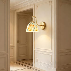 Puede incluir: Lámpara de pared de latón con pantalla de mosaico. La pantalla presenta un patrón de piezas de vidrio de colores azul, naranja y amarillo. La lámpara está montada en una pared crema en un pasillo.
