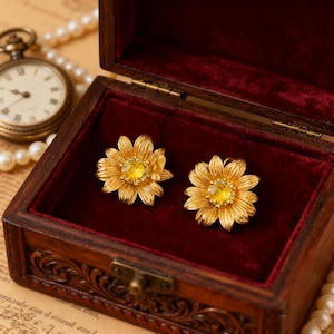 May include: A pair of gold-colored daisy earrings with yellow centers, displayed inside a vintage wooden jewelry box with a red velvet lining. The box is open, revealing the earrings. A pocket watch and pearl necklace are in the background.