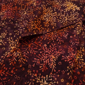 May include: A dark purple fabric featuring a repeating batik-style pattern of orange and yellow snowflake-like shapes. The design includes intricate branching lines and small star-like accents scattered across the deep plum-colored background.