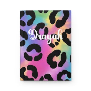 May include: A hardcover journal featuring a vibrant rainbow tie-dye background with a black leopard print pattern overlay. The name Drayah is printed in white cursive script across the center of the front cover of this rectangular notebook.