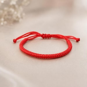 May include: A bright red cord bracelet featuring a tightly woven, braided design. The bracelet includes an adjustable sliding knot closure with two loose cord ends, each finished with a small, simple knot at the very tip of the string.