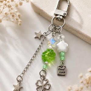 May include: Silver-toned keychain with a lobster clasp, adorned with charms. Features a chain with star pendants, a four-leaf clover, green and white beads, and a small tag that reads "Good Luck".