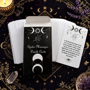 May include: A deck of oracle cards titled Spitir Messages in a black box with white celestial graphics including a sun, moon phases, and stars. One card displays text about personal change, shedding layers, and new beginnings in a clear black font.