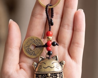 May include: A gold-colored keychain featuring a lucky cat charm with raised paws, a coin, and a gold ring. The charm has a smiling face and the word "福" is visible. The keychain is attached to a brown cord with red and black beads.