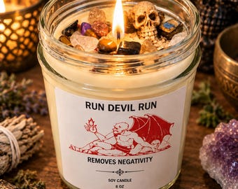 May include: An 8 oz soy candle in a clear glass jar with a lit wick. The wax is topped with various polished gemstones and a small skull figurine. The white label features the text RUN DEVIL RUN, REMOVES NEGATIVITY, and a red illustration of a winged demon.